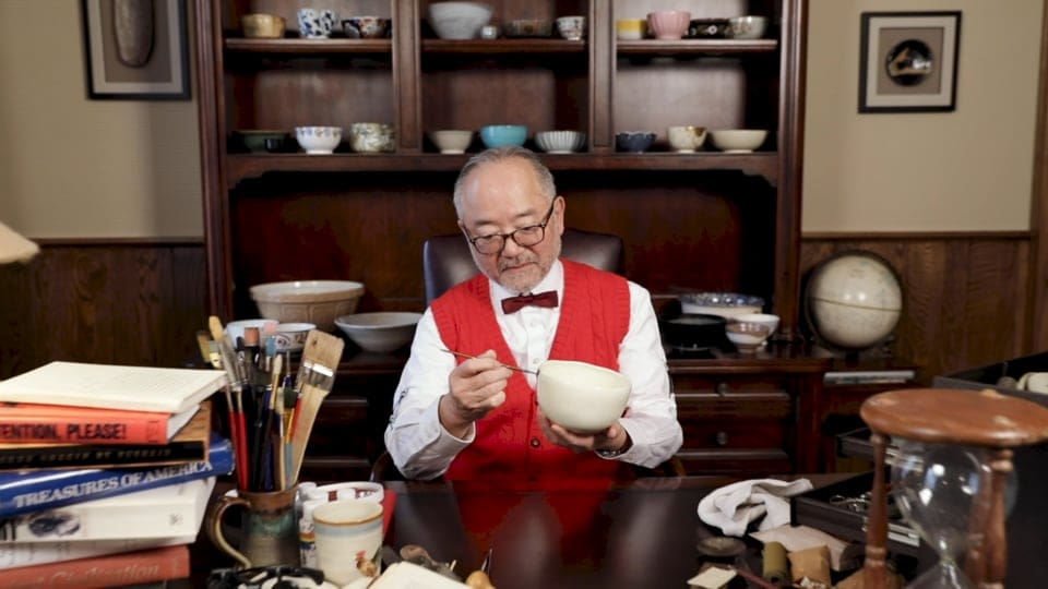 Photo of a daper dressed man painting a ceramic bowl at a desk, with a cabinet full of ceramic bowls behind him.