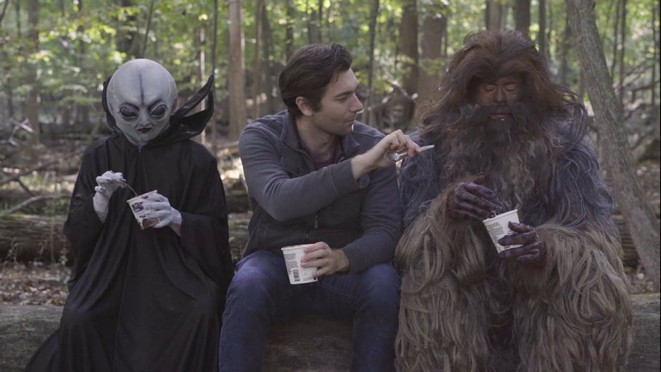An alien in a black cloak, a human man, and a sasquatch sit on a log in a forest together and eat cups of noodles. The man offers a fork to the sasquatch.