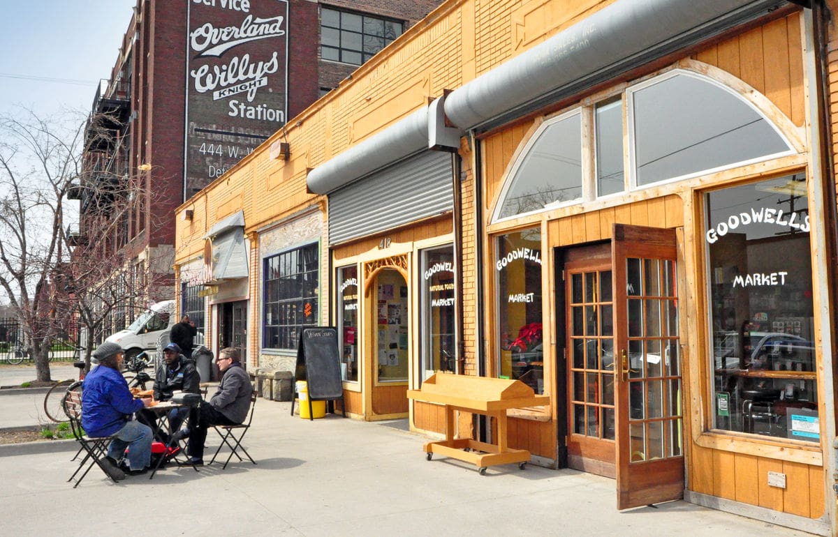 wide shot photo of the exterior of a market in Detroit. The building is yellow brick and light wood, with white text painted in the window that says "Goodwells Market".