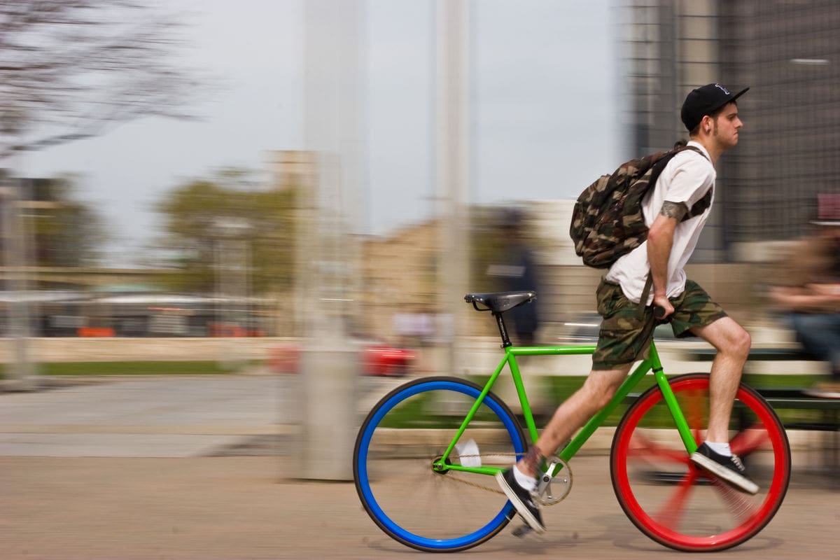 Motion blurred photo of a man riding his bike down an urban street. The bike has a green frame, a red front wheel and blue back wheel.