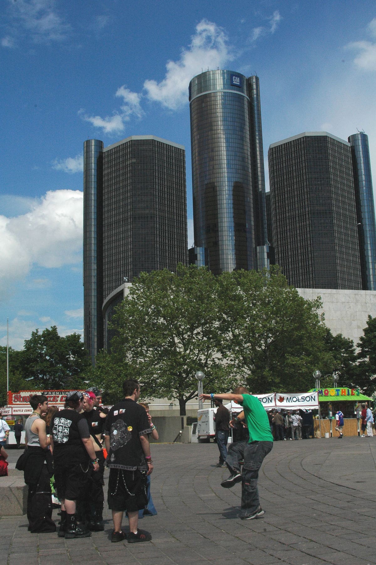 Photo of a crowd at an outdoor fair in Detroit on a sunny day. In the distance are three high rise buildings.