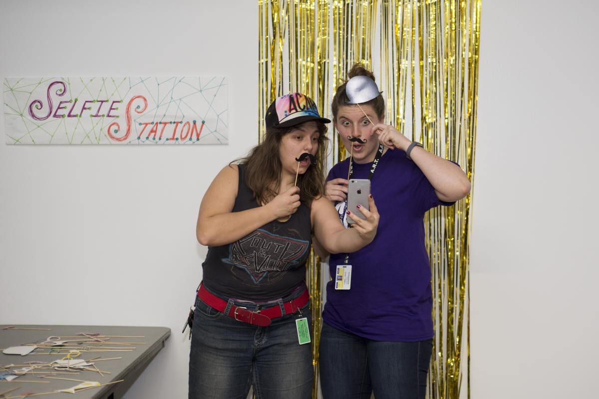 Photo of two CCS students making silly faces and holding up photo booth props up to their face while taking a photo. They are standing next to a sign that says "Selfie Station" posted above a table full of more photo booth props.