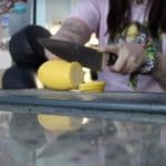 Close-up wide shot photo of someone cutting a yellow squash with a huge knife on a black cutting board. The table is reflective white marble, and a household kitchen is visible in the background.