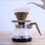 Closeup photo of someone pouring water from a white ceramic kettle into a coffee filter full of grounds. The filter is on top of an espresso kettle already partially filled with coffee. Pictured on a wooden table against a white wall.