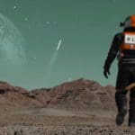 Wide-shot photo of an astronaut in a black suit with a grey and orange helmet and backpack facing away from the camera and walking across the brown rocky planet terrain. The sky is blue-green, and in the distance is a shooting star and the faint image of a giant planet.