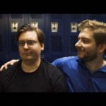 Photo of two men in a locker room. One man has his arm around the other, looking at him very smugly. The other man looks annoyed.