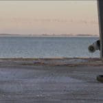 Wide-shot photo of the ocean coast at sundown. In the foreground is the leg of someone in black pants and sneakers holding a skateboard.