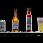 Advertisement that shows a beer can, two beer bottles, and glass of beer arranged on a tray of ice on a black background with dramatic lighting. The brand is Budweiser.