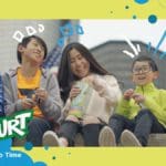 Photo of five smiling children sitting on the side of the road and enjoying Go-Gurt products. The children are decorated with yellow, blue, and white details drawn on top of them. In the bottom left corner is a Go-Gurt logo.
