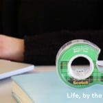 Closeup photo of someone in a black sweater working on a laptop in a library. To the right is a roll of scotch tape with green packaging. Below it is white text that says "Life, by the roll."