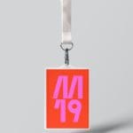 Closeup photo of a badge on a white lanyard on a white background. The badge is neon orange with neon text that says "M 19"