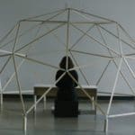 Photo of a woman in all black sitting at a white wooden table, facing away from the camera. She is under a large wooden geometric dome sculpture.