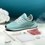 Photo of a white Adidas sneaker with decorative teal mesh on top. There is various plastic trash arranged around the sneaker to represent that it was made of recycled material.