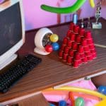 Photo of a wooden desk in a pink room with a bunch of knick knacks. The objects include a tower of small red plastic cups, a desk fan with balloons attached, and a small old-school computer.