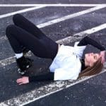 A women in a white hoodie, black leggings, and yellow sparkly eye makeup poses on the ground on a parking space.