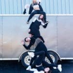 Five people standing on top of each other in various black and white outfits pose in front of a silver trailer.