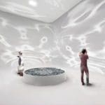 Photo of two people in a white room with giant water ripple reflections on the wall. A pool of water in the center of the room provides the reflections.