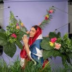 Photo of a woman wearing the Puerto Rican flag posed in front of a lilac sheet surrounded by exotic plants.