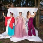 Photo of three women posing in elaborate dresses in a background against a white sheet hung up on a metal frame.