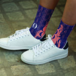 Up close photo of someone wearing white sneakers and pink and dark blue socks. The socks have pink text and images on them. The ground is paved cobblestones.