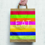 Photo of a hand holding a paper back with red, green, yellow, dark blue, and pink stripes. In the center is white text that says "Eat". The background is a white paved wall.