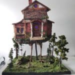 Painted sculpture of a raggedy red wooden cabin balanced on two long chicken legs. The house is covered in moss, and it is standing in a mossy and tree covered forest display.