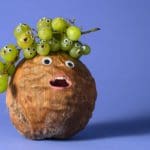 Coconut with a branch of grapes on top in front of a blue background. All of the fruit has googly eyes and a human mouth.