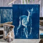 Blue painting of a deer in front of various decorative stumps and sticks of wood.