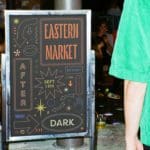 Picture of a chalkboard advertising the Eastern Market after dark on September nineteenth. All the text is brightly colored and stylized. The board is decorated with various arrows, hands, lips, and geometric shapes. The color scheme is yellow, orange, pink, and dark blue.