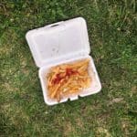 Overhead shot of french fries with ketchup in an open Styrofoam box sitting in grass.