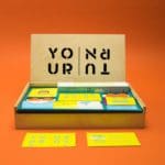 Photo of a wooden box with an open lid that has black text which reads "Your Turn". Pictured on an orange background. The box is full of yellow, blue, and white business cards and other products.