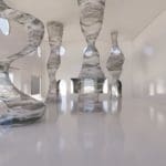 Photo of five tall, wavy, grey, semi-transparent sculptures in a white art exhibition room.
