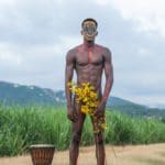 Photo of a man in red and yellow body paint holding yellow flowers standing in a field. To his left is a traditional drum.