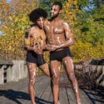 Photo of two men in black shorts and white body paint embracing on a wooden bridge with yellow and green trees in the background.