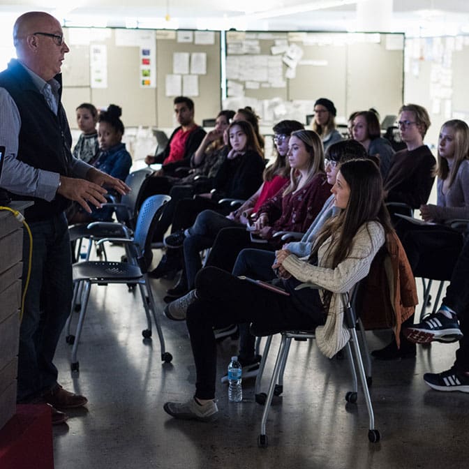 Classroom full of students sit in chairs and listen to a professor give a speech.