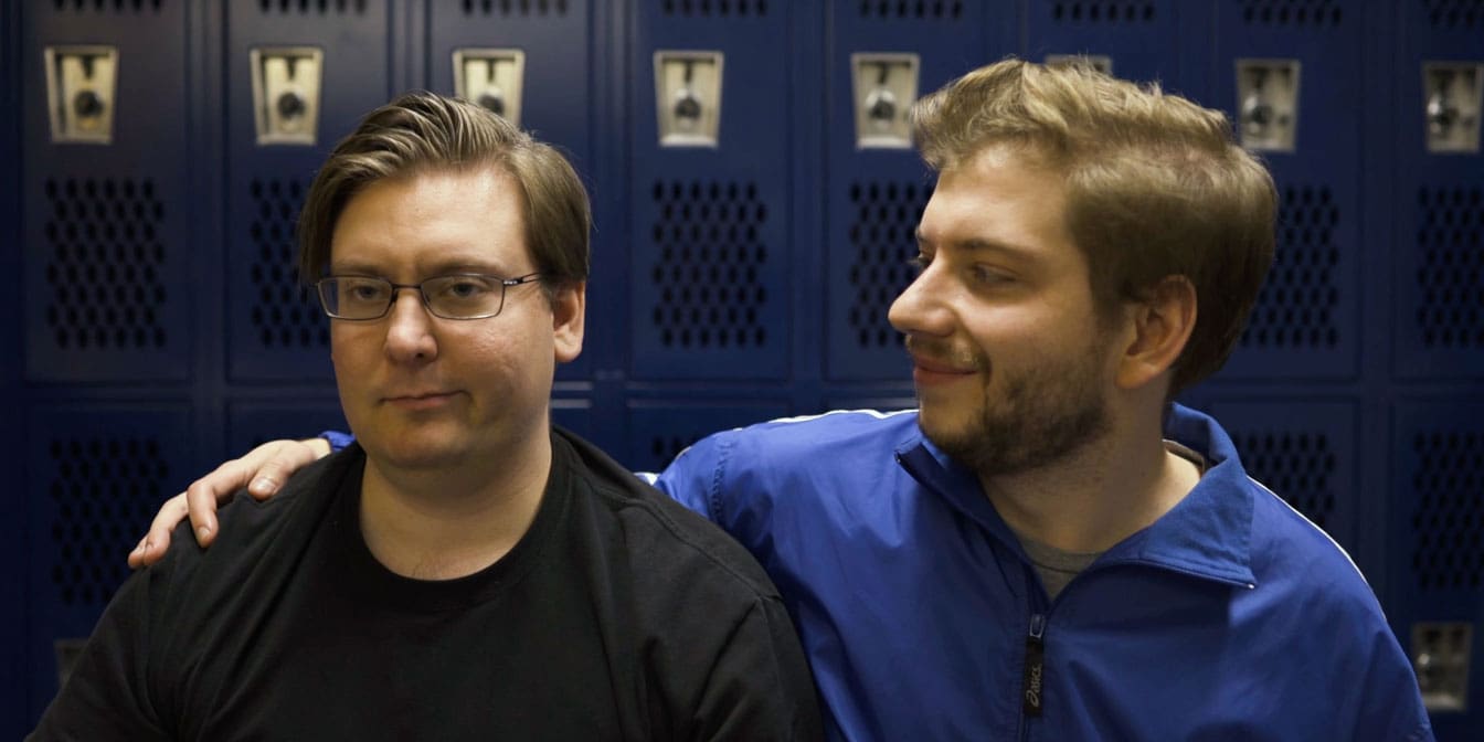 Photo of two men in a locker room, where one man looks annoyed, while the other man has his arm around the first man and smirks in his direction.