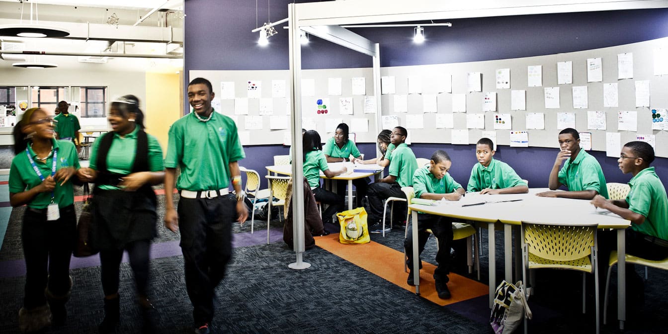 A group of students in school uniforms sittig as tables and walking down the hall in a school setting. Their school shirts are green.