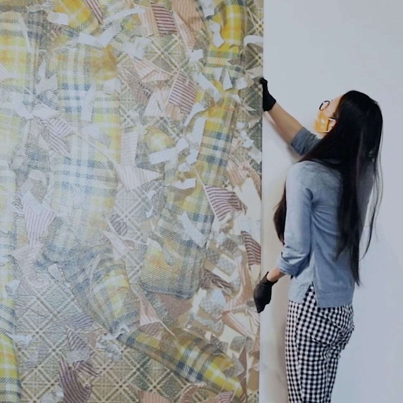 An artist installs her giant art piece on the wall. The piece is an abstract collage of neutral colored plaid patterns.