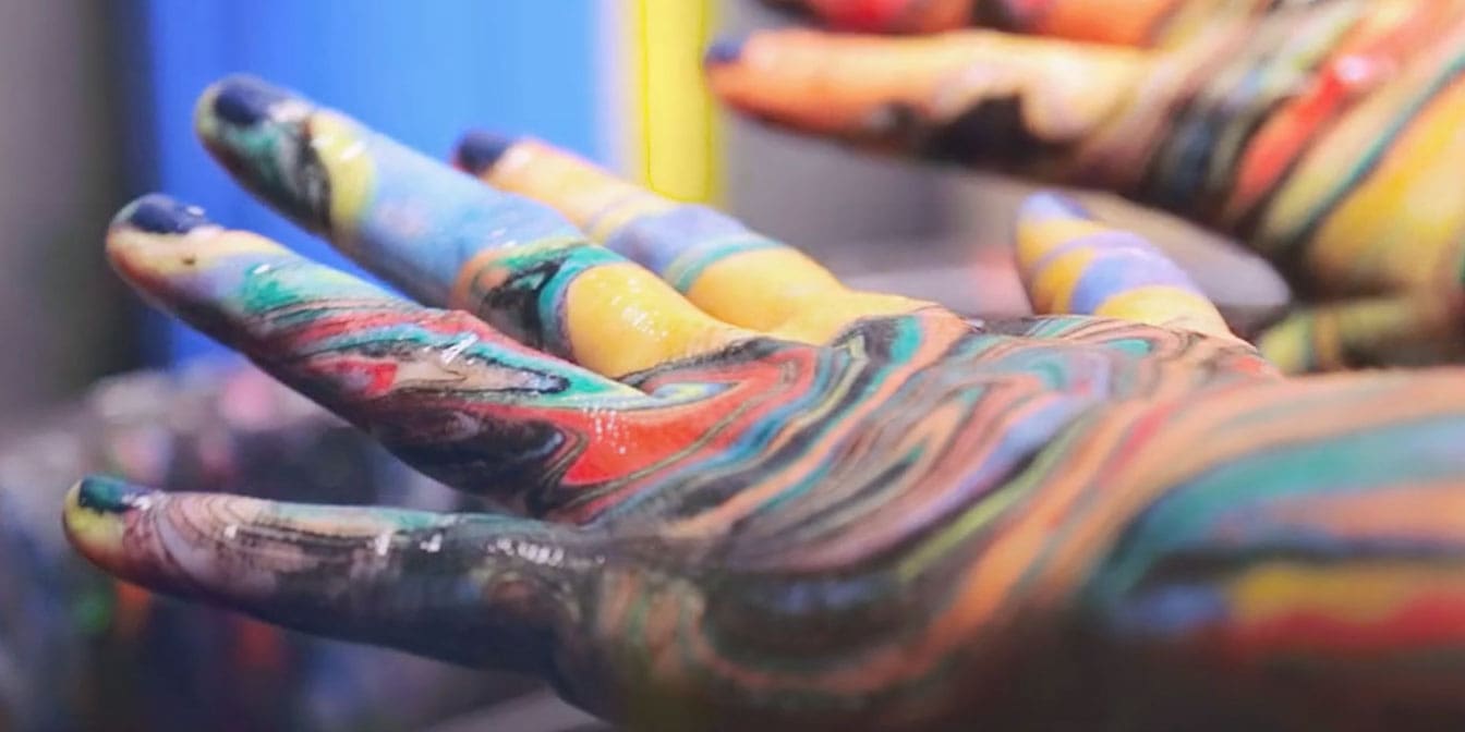 Close up photo of two hands that are covered in swirls of red, black, yellow, blue, orange and pink paint.