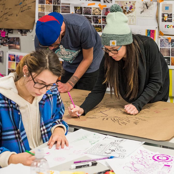 communication design projects Three people draw on large sheets of paper together in a classroom