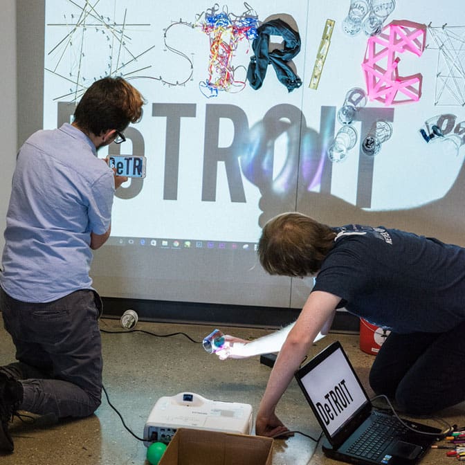 Two people work on a projector and a laptop in front of a wall with a graphic design display projected onto it.