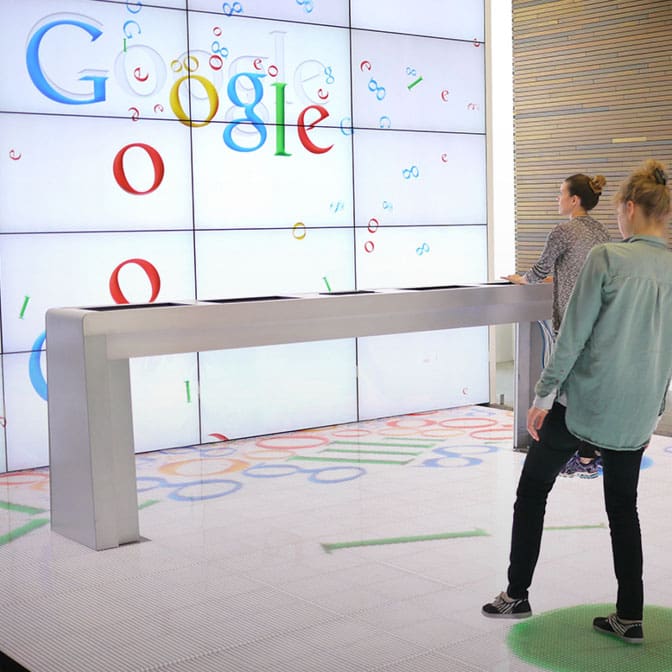 Photo of two people in front of a large wall full of screens that display the Google logo