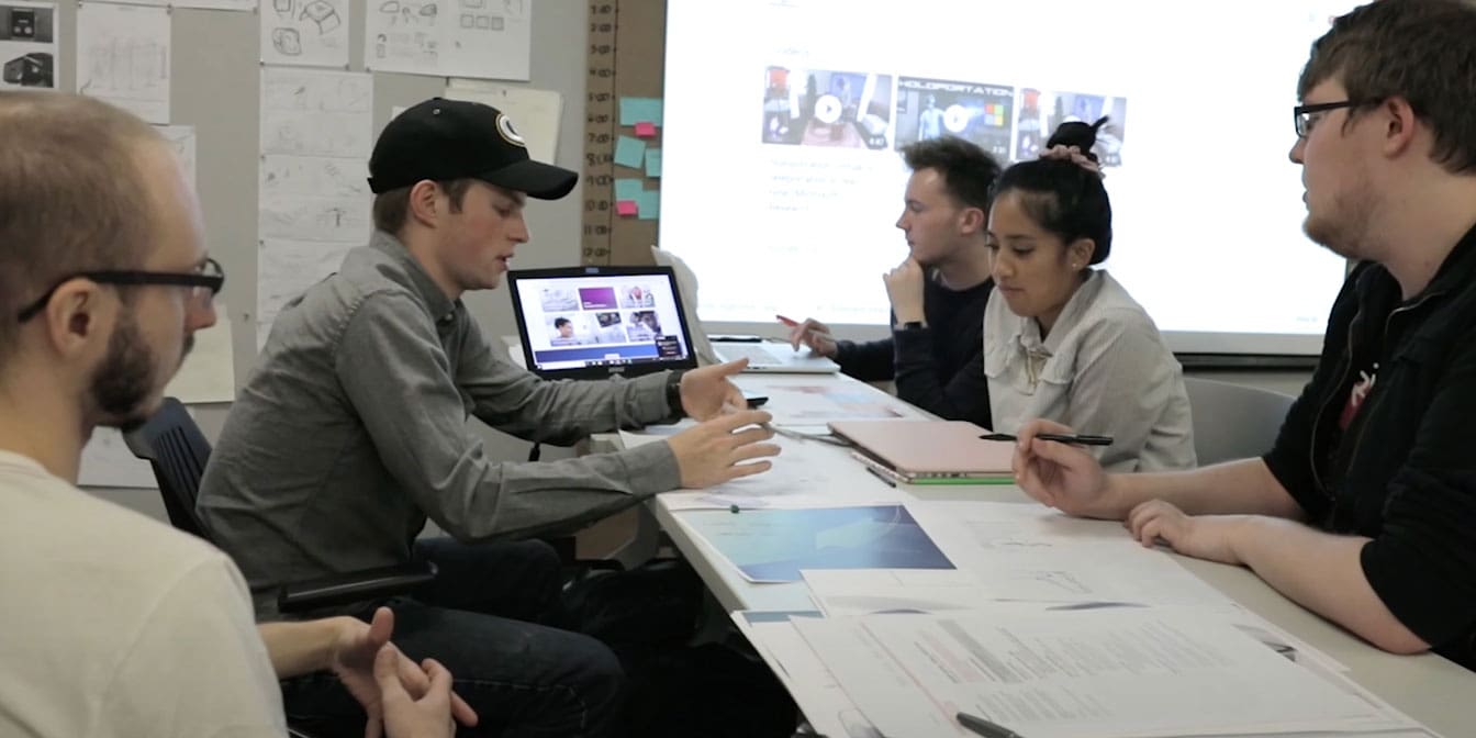 communication design projects Photo of five people having a discussion in front of a laptop in a meeting room