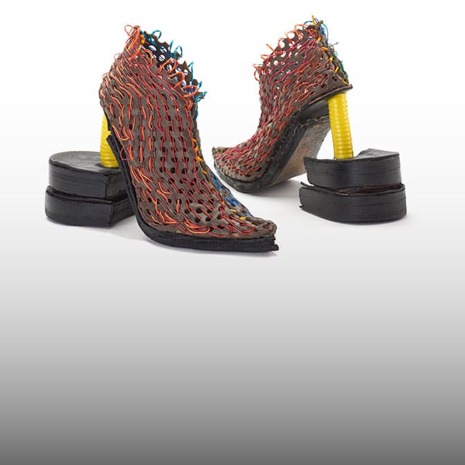 Photo of a pair of futuristic concept high heels. The heels are yellow with large black rubber soles. The shoe itself is grey with red and orange string weaving in and out all over the shoe.