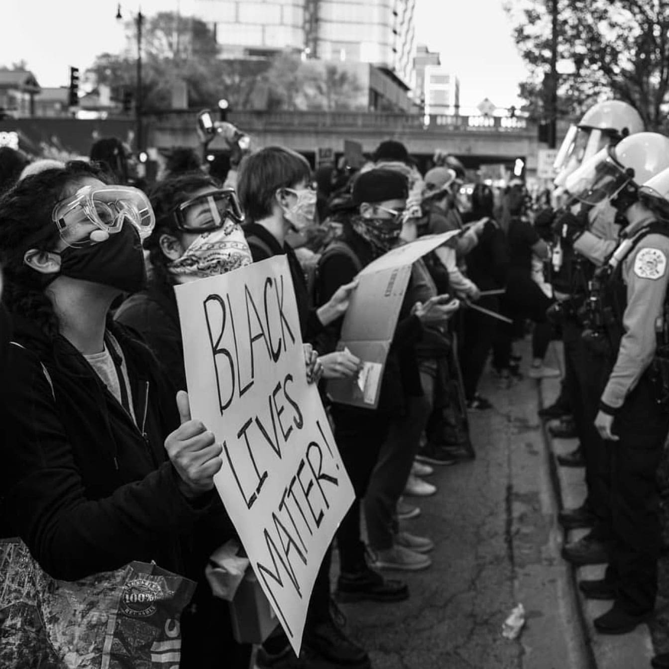 Black and white photo of protestors facing a line of policemen. One protestor holds a sign that says "Black Lives Matter!"