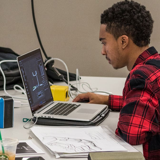 Photo of a man working on his laptop with his sketchbook in a classroom