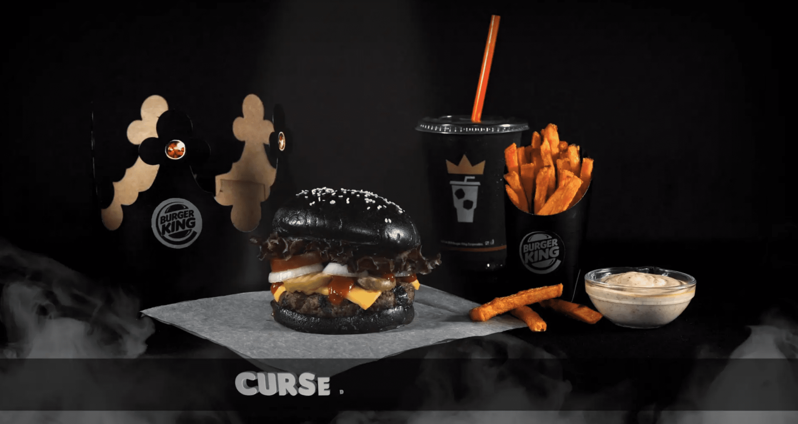 Advertisement for Burger King. Shows a paper crown, cup of fried, and soda cup, which are all black. In the center is a burger with black buns. The background is also black.