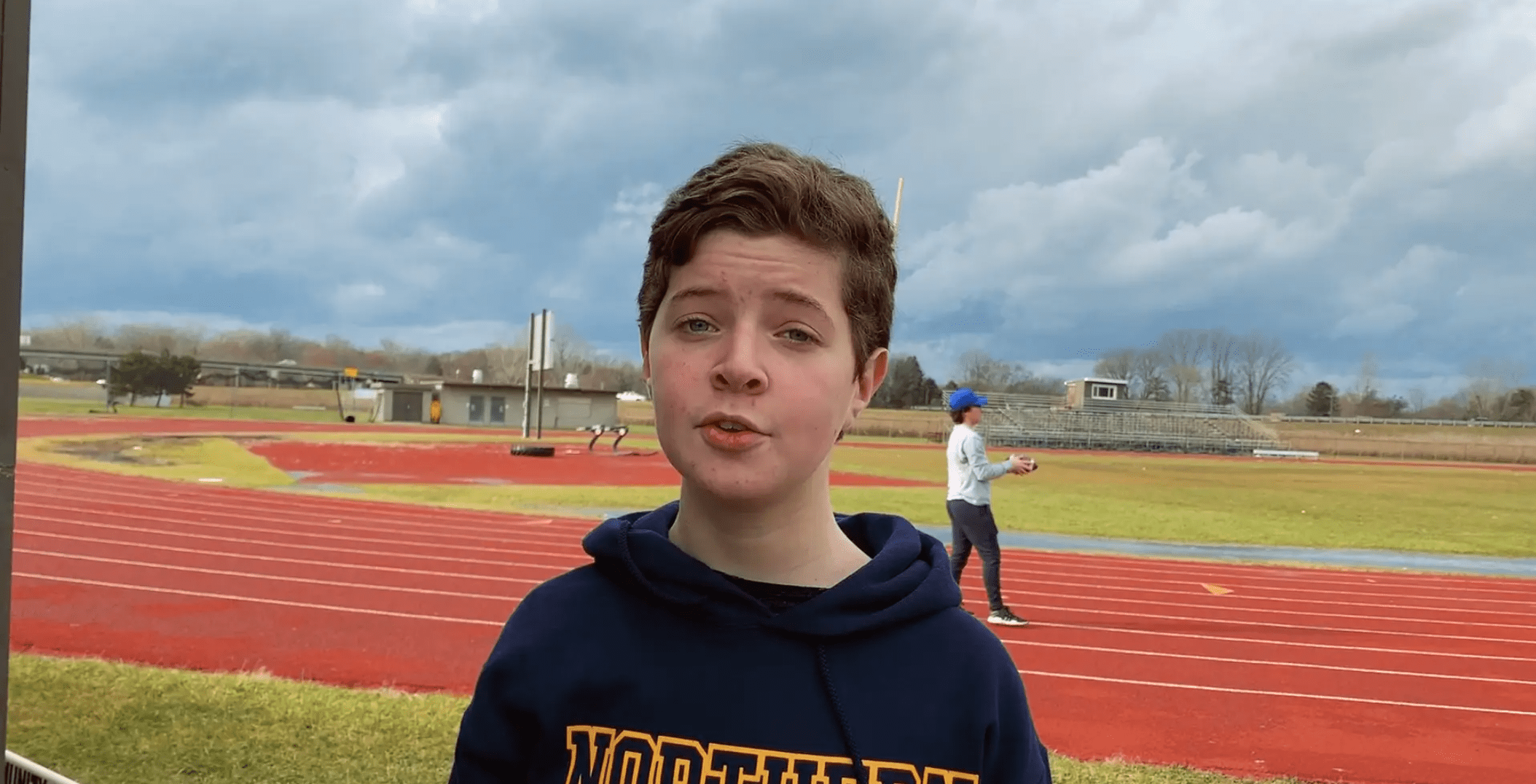 Photo of a young boy in a navy blue hoodie speaking. He is standing in front of a high school running track.