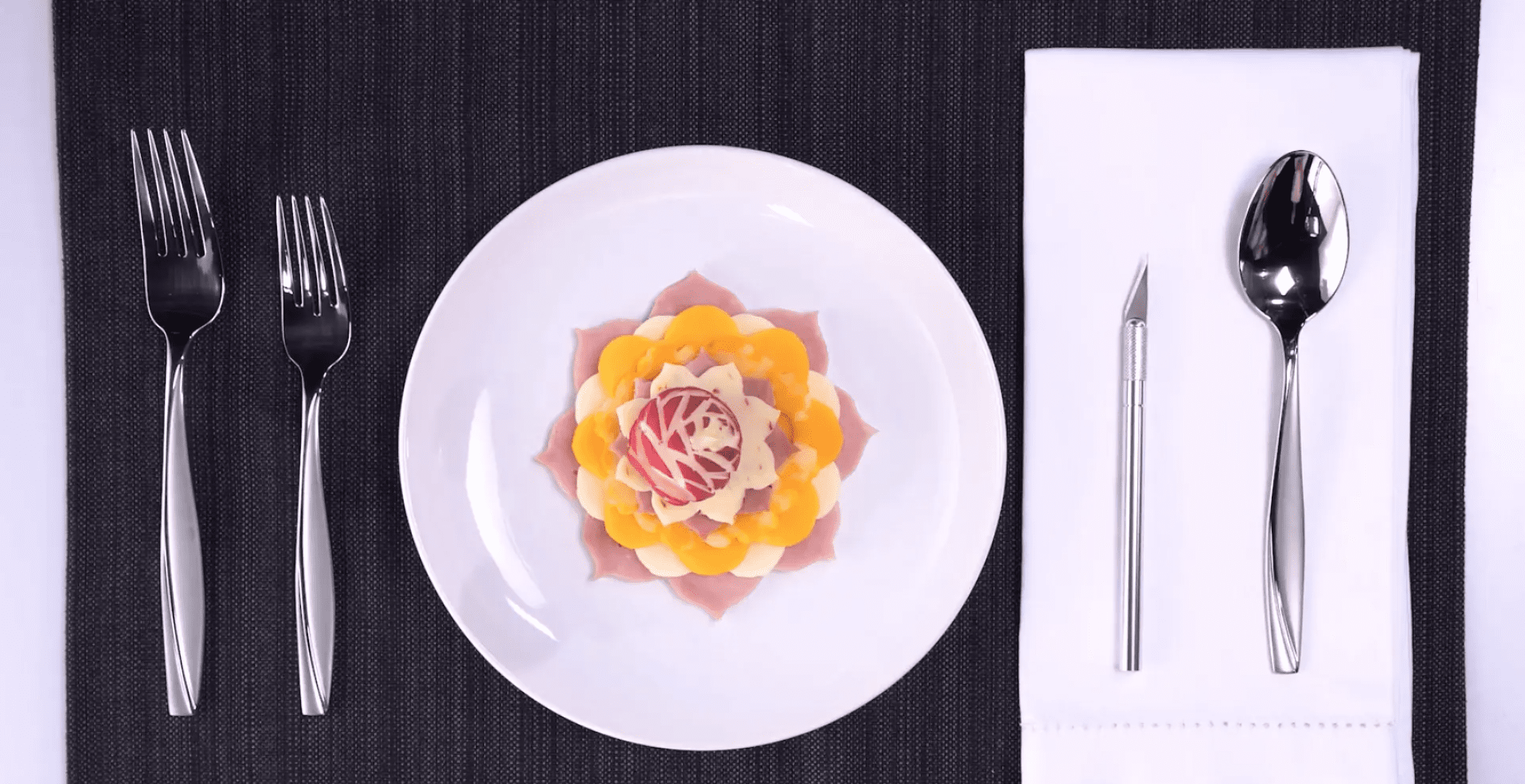 Photo of a white plate on a black wood table with silver utensils on either side, and a white napkin to the right. On the plate is cheese, ham, tomatoes, and other foods arranged to look like a flower.