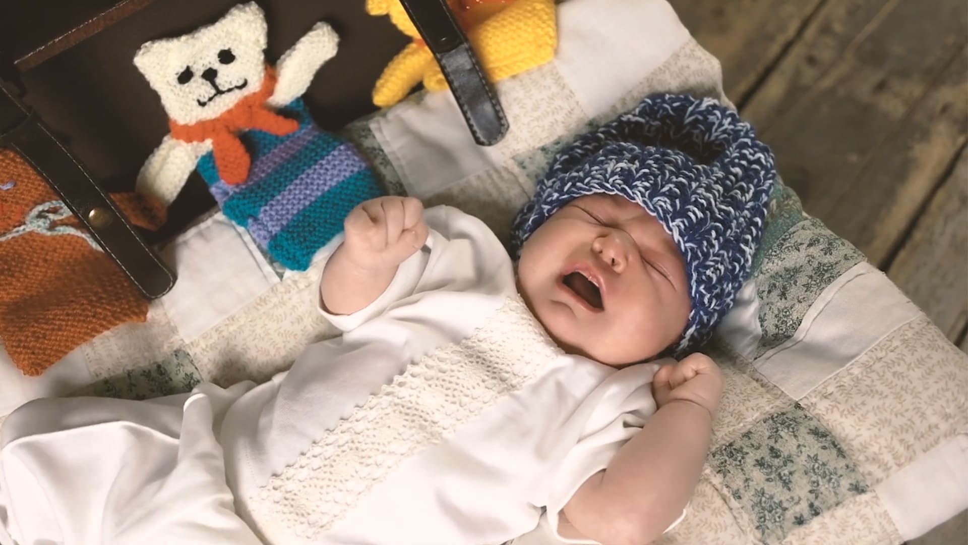A screengrab of an advertisement depicting a crying baby dressed in white and a blue knitted hat laying on a white patch blanket. The view is from above.